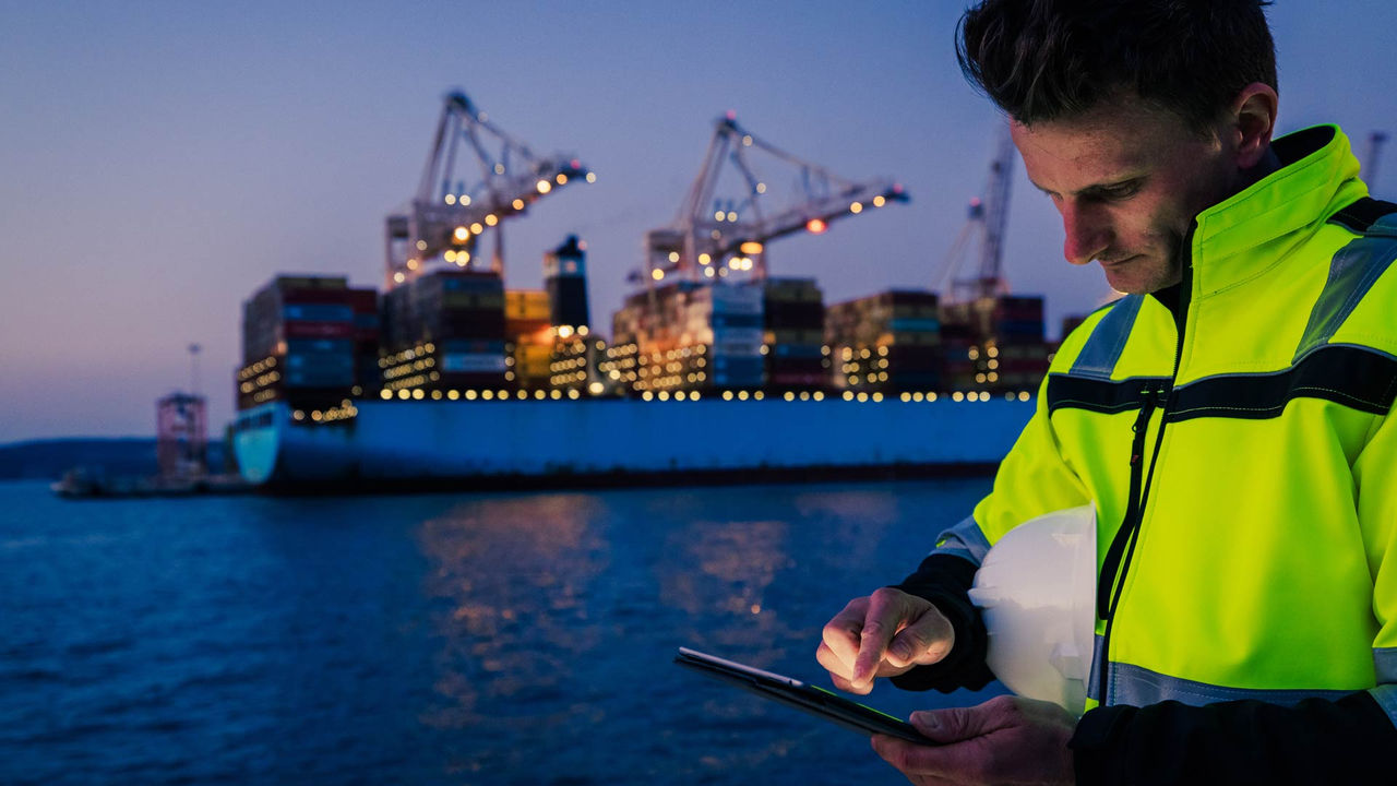 Port worker uses a tablet beside container ships and cranes, illustrating global supply chain operations. 